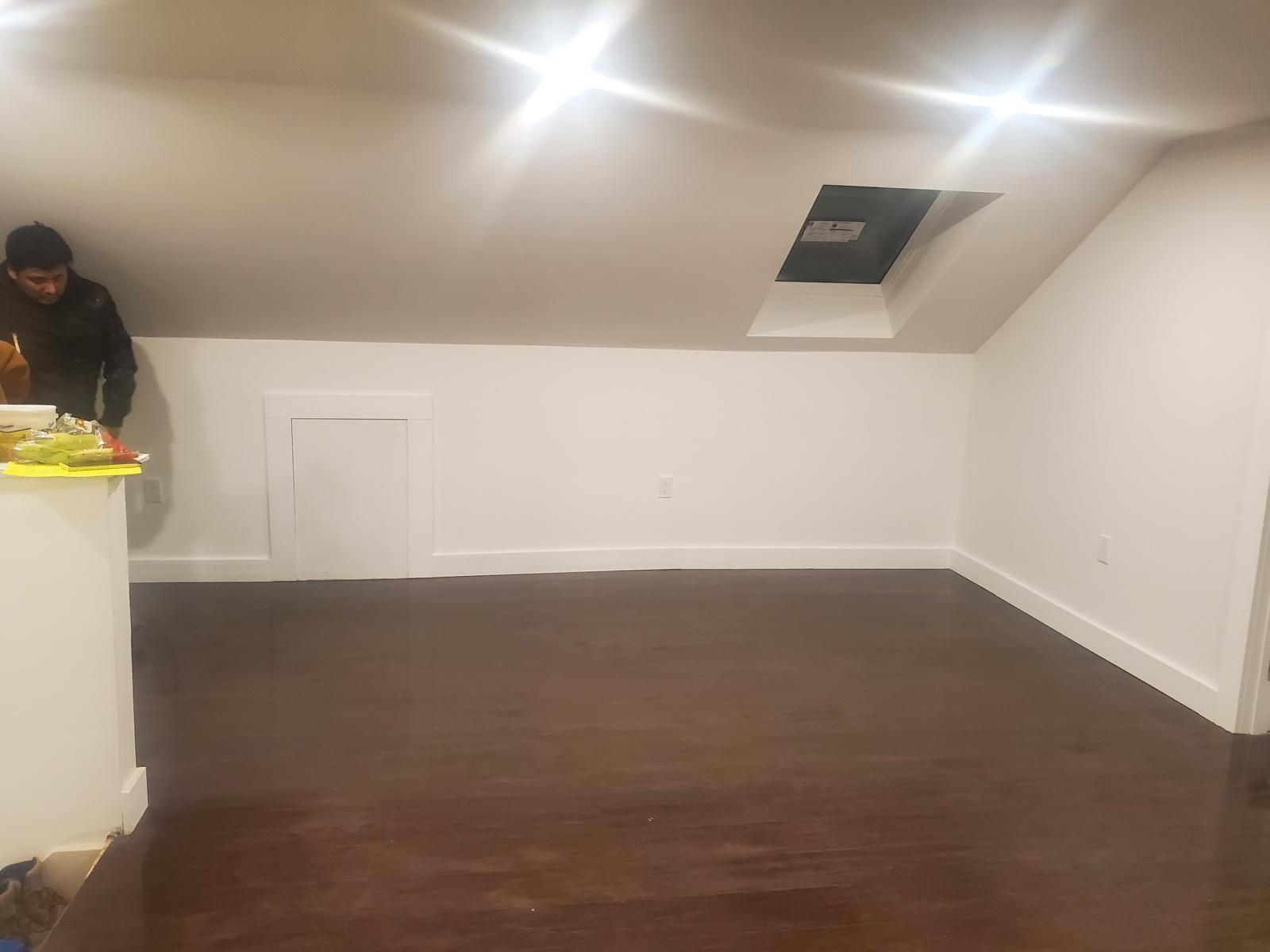 Empty room with dark wood floor, white walls, and a man partly visible at a counter on the left.