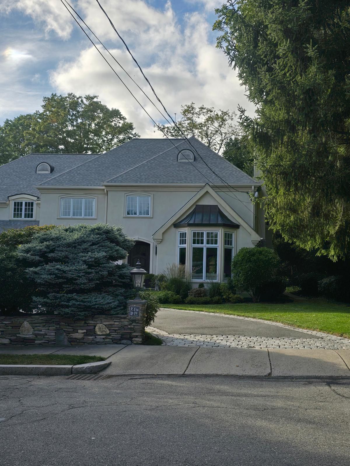 Suburban house with gray roof, front garden, driveway, and tall trees under a cloudy sky