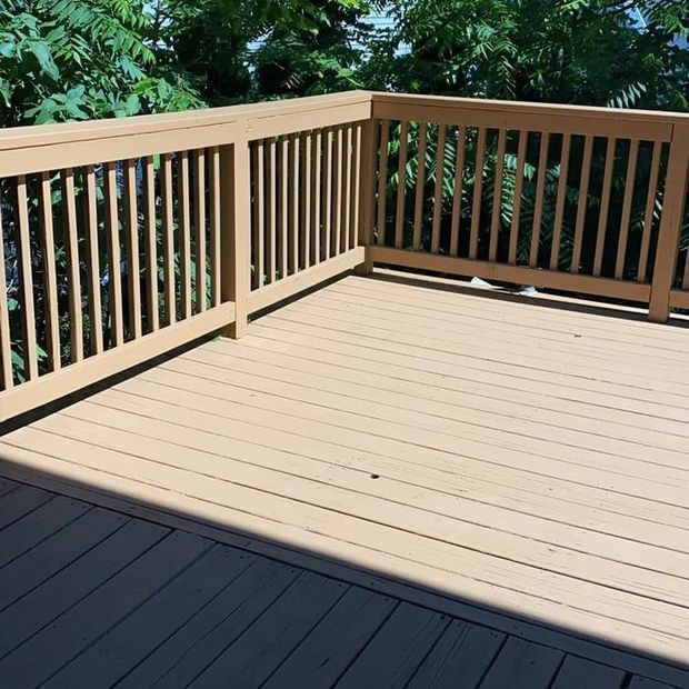 Sunlit wooden deck with tan railing and dark gray flooring, bordered by green trees