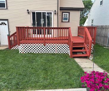 Red wooden backyard deck with stairs, white lattice skirting, and a concrete path beside a lawn and flowers