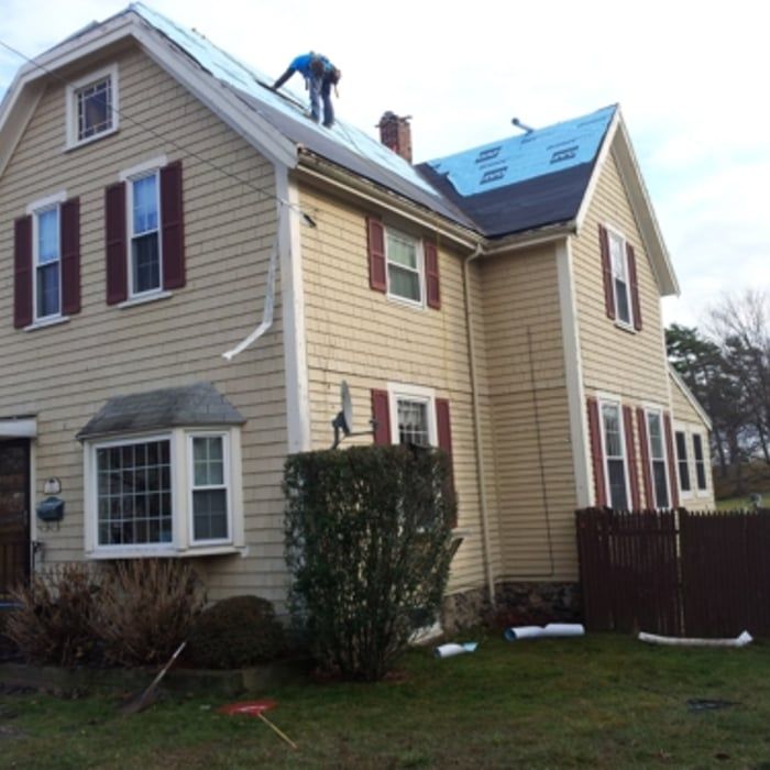 Worker on a roof of a beige two-story house with blue shingles and white-trim windows