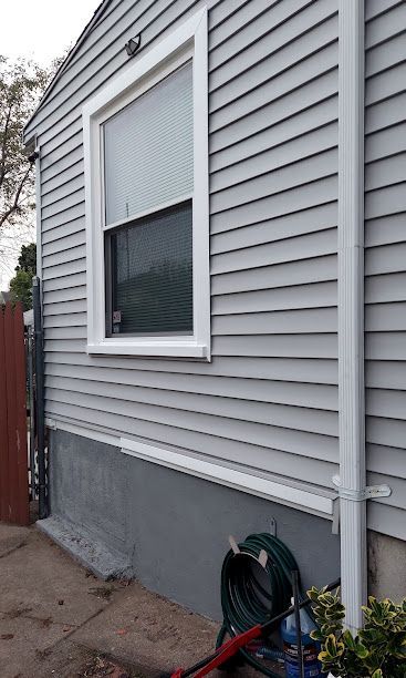 Side of a gray house with a white-trimmed window, concrete foundation, and a garden hose on the ground.