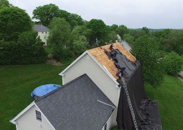 Firefighters work on a roof damaged by fire beside a house surrounded by trees.
