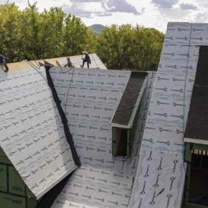 Workers installing gray roofing underlayment on steep roof sections, with trees in the background.