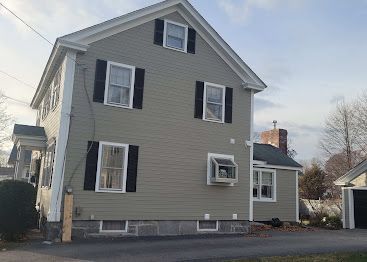 Gray two-story house with black shutters and white trim on a quiet street under a cloudy sky