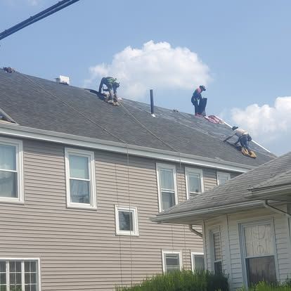 Two workers on a gray shingled roof, repairing shingles on a house under a blue sky.