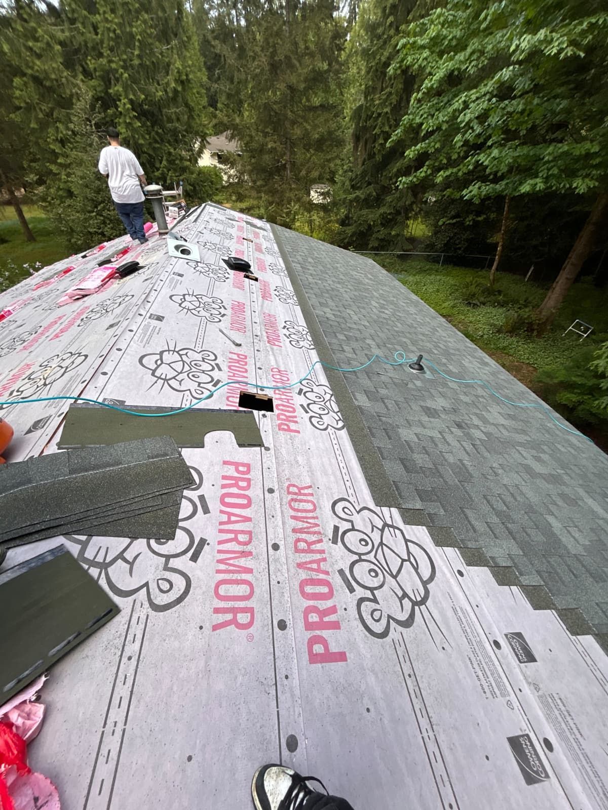 Workers installing gray roofing underlayment on a steep roof surrounded by trees