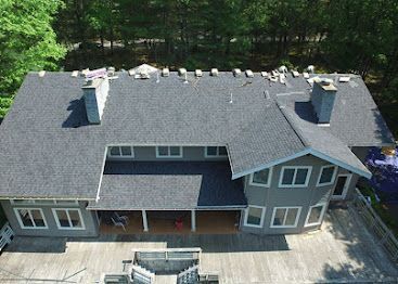 Aerial view of a gray roofed house with dormers and a front porch surrounded by trees
