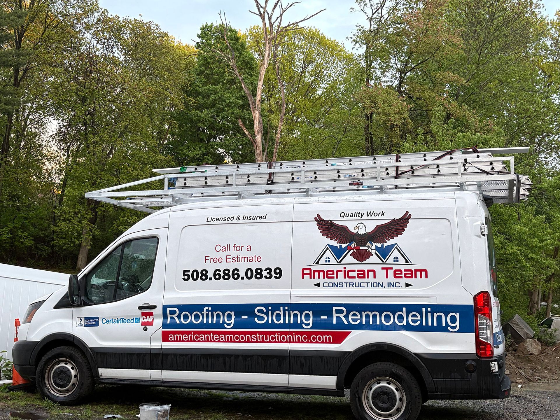 White American Team roofing van with ladder and company logo parked outdoors near trees.