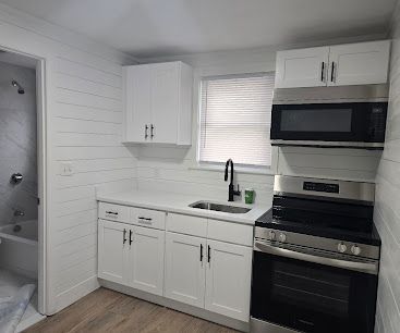 Small white kitchen with sink, cabinets, stove, microwave, and a window with blinds.