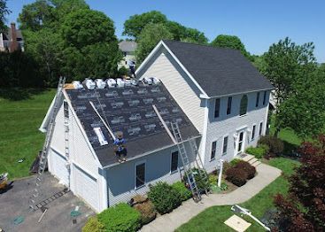 Aerial view of a white house with a dark roof and solar panels, surrounded by trees and lawn