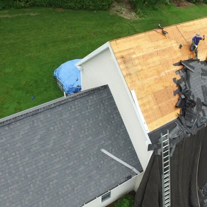 Aerial view of roof replacement with workers on a house and roofing materials piled on the right.