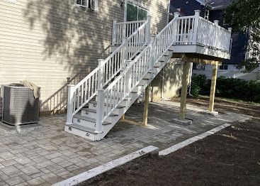 White exterior staircase and deck beside a beige house, with air-conditioning unit and paved walkway.