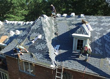 Roofers working on a steep house roof, laying dark underlayment around a dormer and chimney.