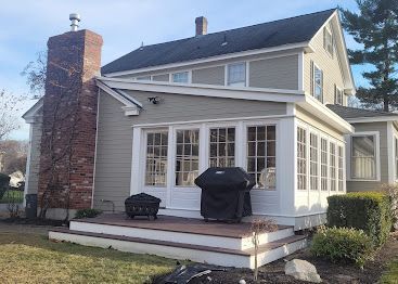 Back porch of a gray house with white trim, windows, and a covered grill on the deck.
