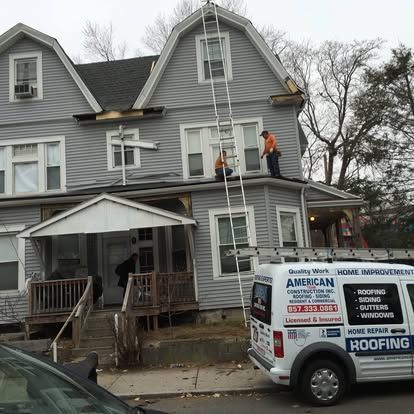 Workers on a gray house roof beside a white roofing company truck in a residential street