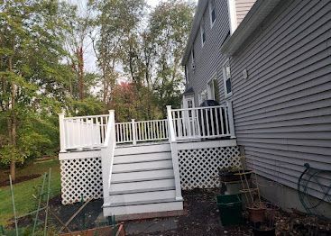 White wooden deck stairs and railing beside a gray house, with a lattice skirt and backyard trees.