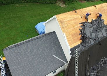 Aerial view of a roof being torn off, with workers on a yellow plywood section and debris on the edge.
