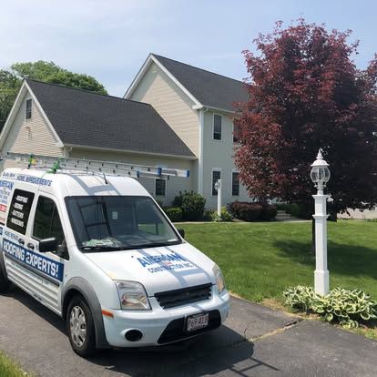 White home security van parked in a driveway beside a beige house with a red tree and lawn post.