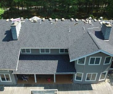 Roofers installing black shingles on a steep house roof beside a white-sided home, with ladders and trees nearby