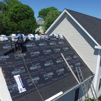 Roofers installing black roofing underlayment on a steep house roof, with ladders and trees nearby