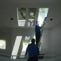 Two men working indoors, one on a ladder reaching towards skylights in a white ceiling.