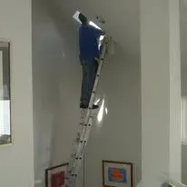 Person on a tall ladder reaching towards the ceiling; inside a white room, near artwork.