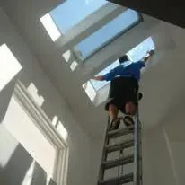 Man on a ladder cleaning a skylight. He is wearing a blue shirt and black pants. Indoors.