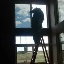 Person on a ladder installing window film inside a building with a view of sky and landscape.
