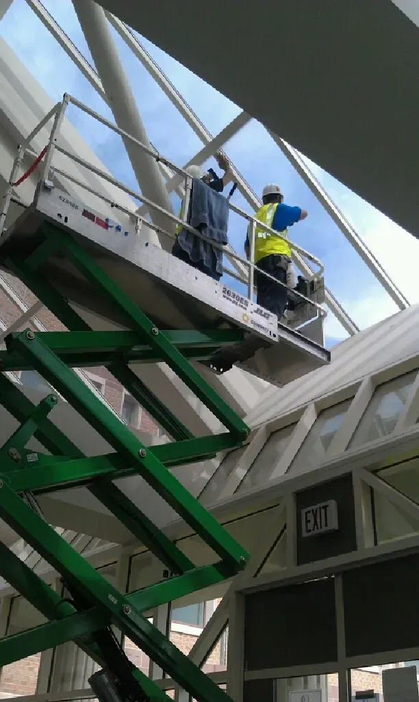 Two workers on a green scissor lift, cleaning a glass skylight in a building.