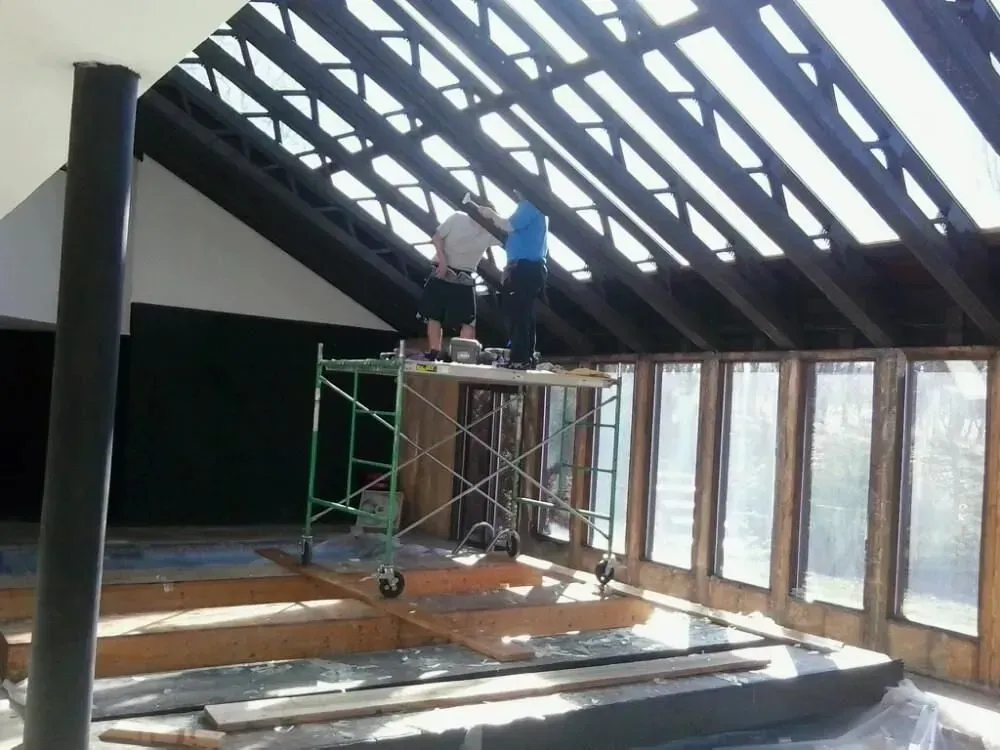 Two people on scaffolding work on a sloped roof with wooden beams and glass windows.