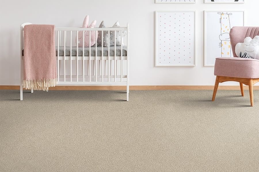 A baby's nursery with a white crib, pink blanket, and tan carpet. A pink chair and artwork complete the room.