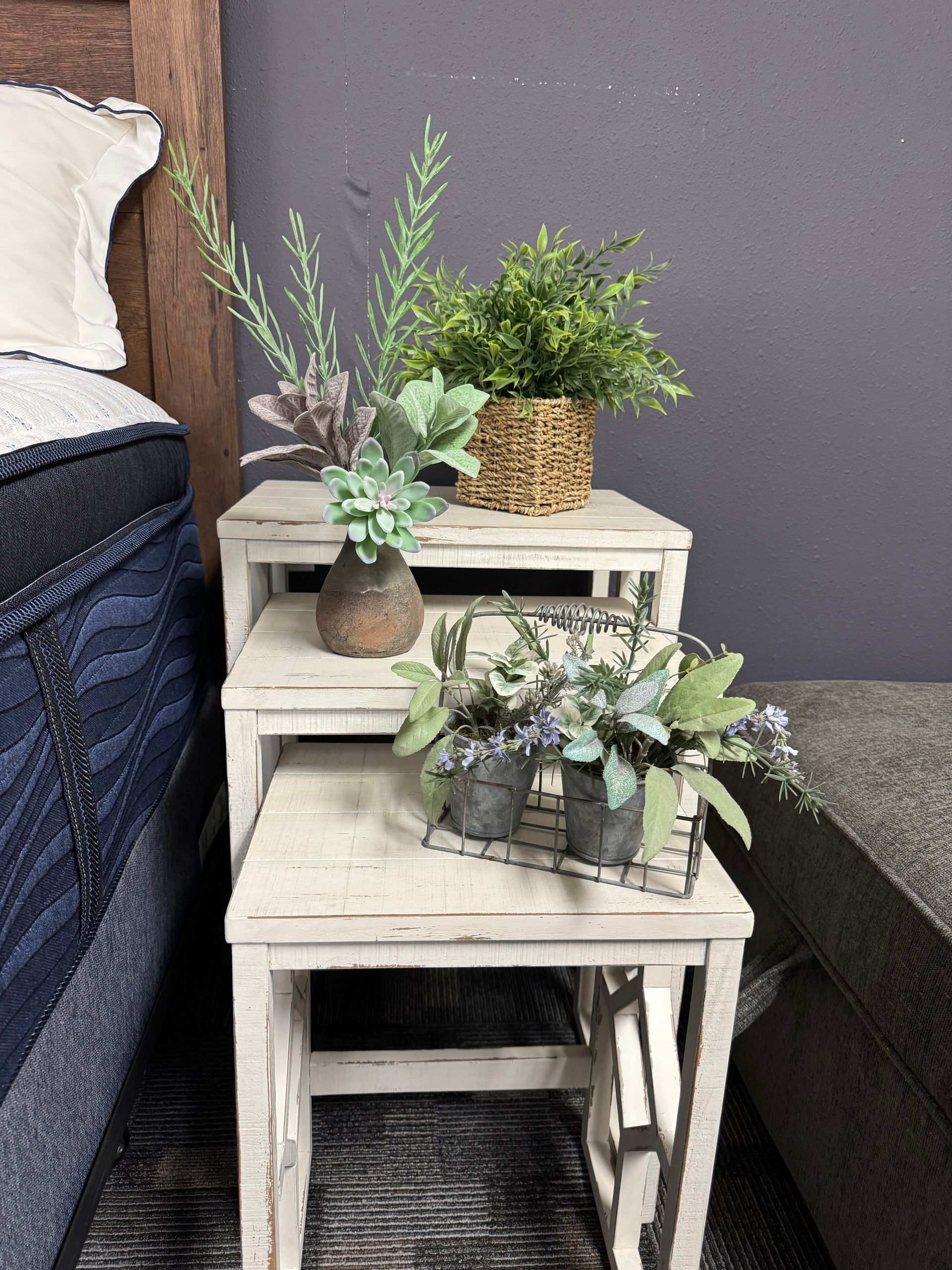 Three-tiered white wooden shelf with plants and decor next to a bed.