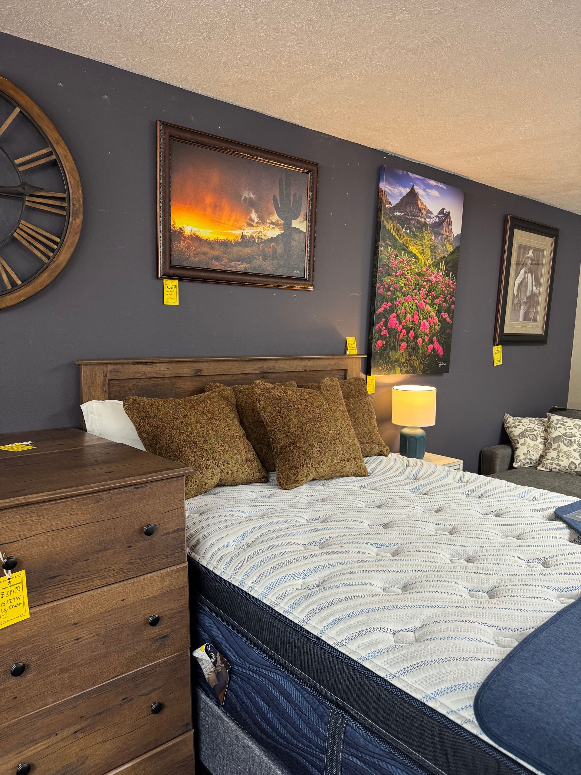 Bedroom furniture display, bed with pillows, dresser, and wall art against a dark gray wall.