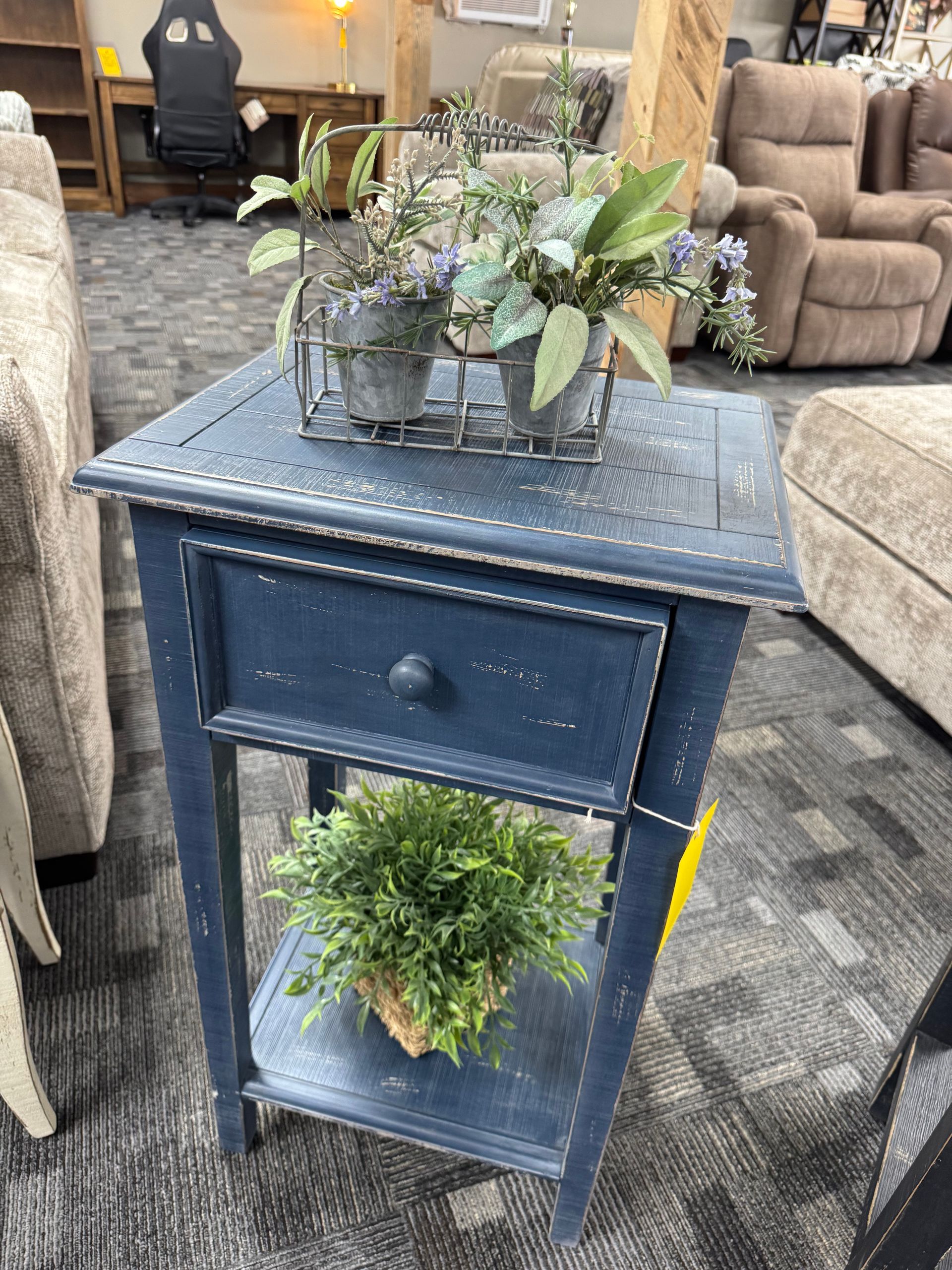 Dark blue end table with drawer and shelf, with plants on top and below, inside a furniture store.