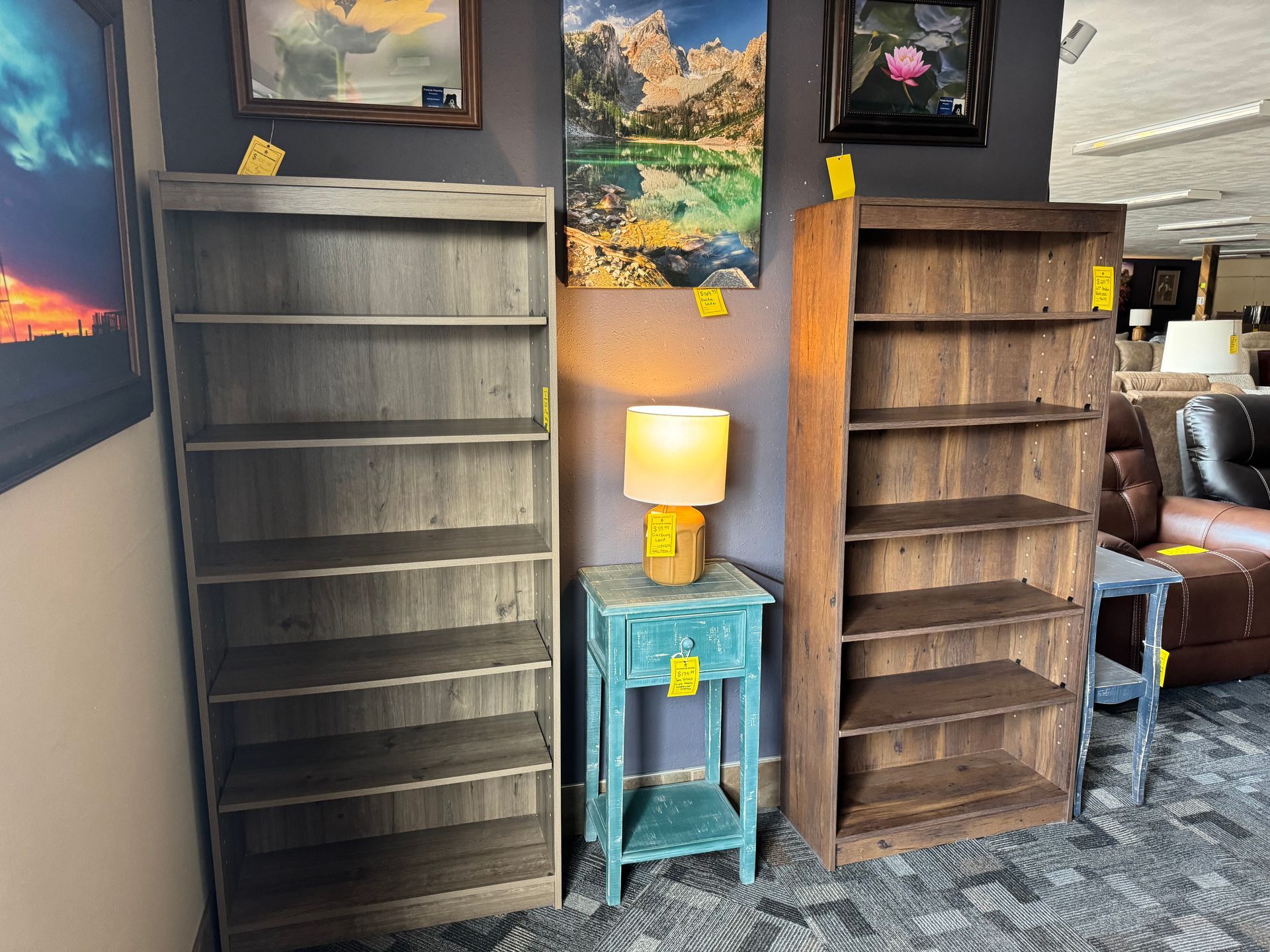 Three wooden bookshelves, a small blue table with a lamp, and wall art in a room.
