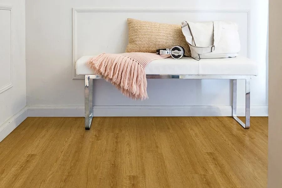 A bench with pillows and a blanket sits against a white wall. The floor has wood-look planks.