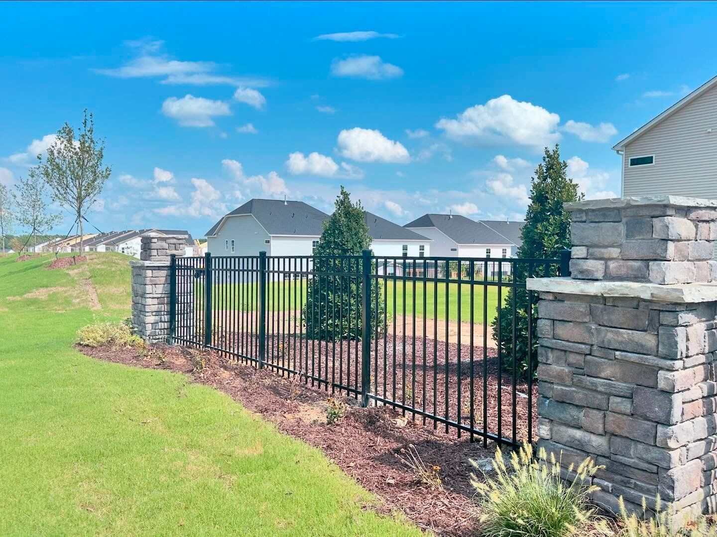 A metal fence surrounds a lush green field with houses in the background.