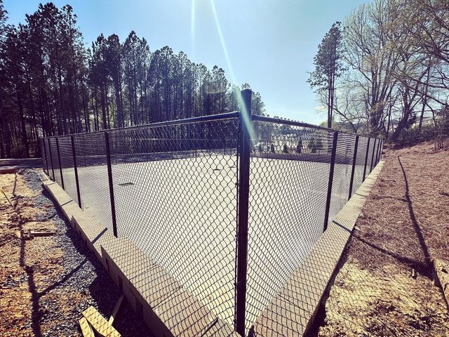 A chain link fence surrounds a tennis court with trees in the background