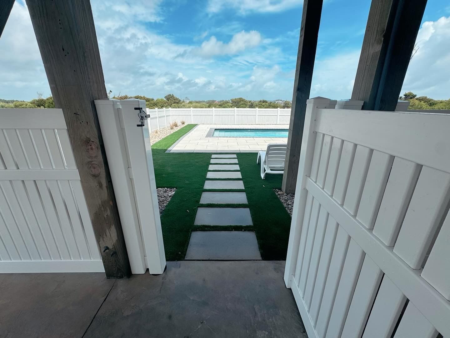 A white gate is open to a walkway leading to a swimming pool.