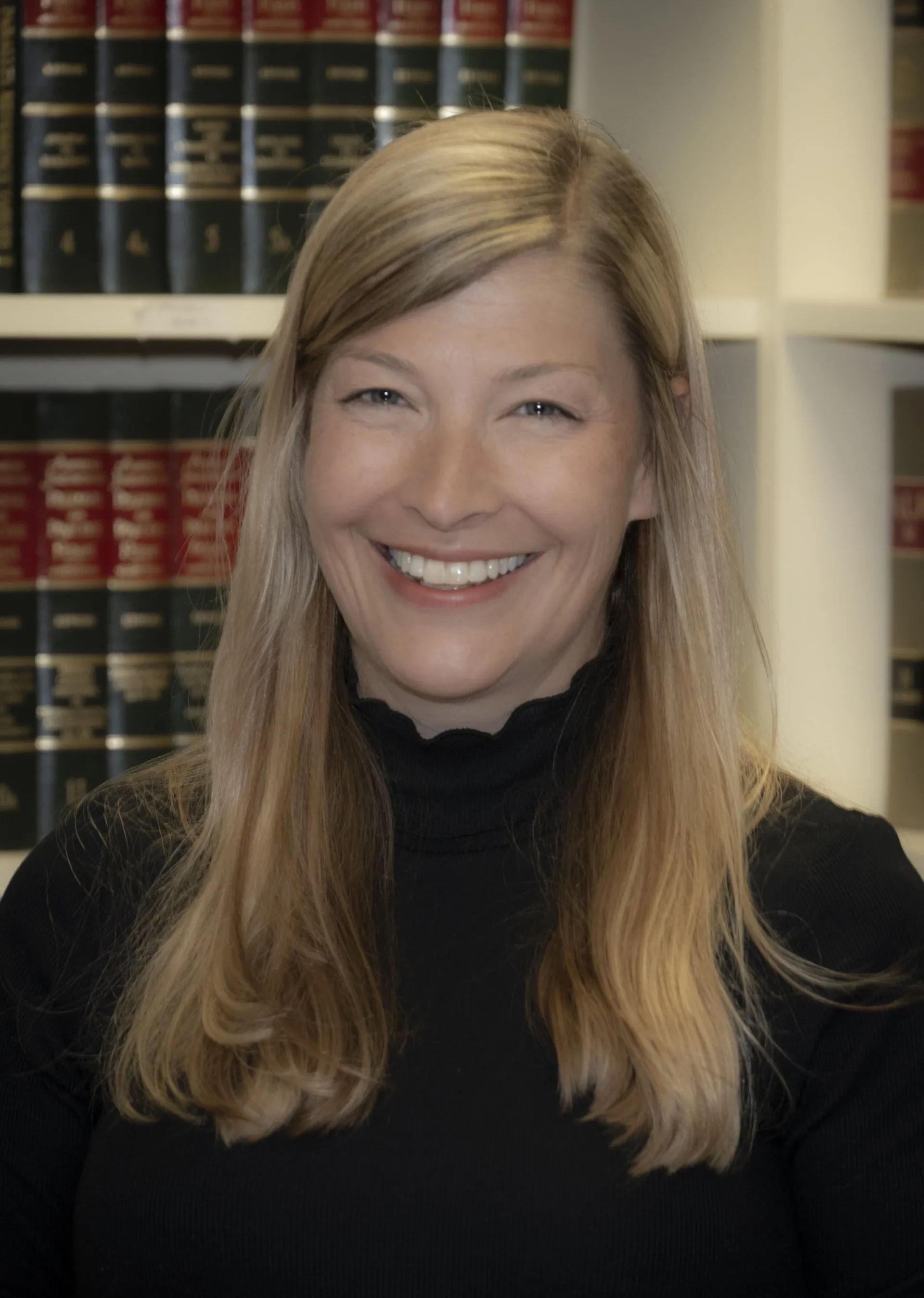 Woman with blonde hair smiles in front of a bookcase. She wears a black sweater.