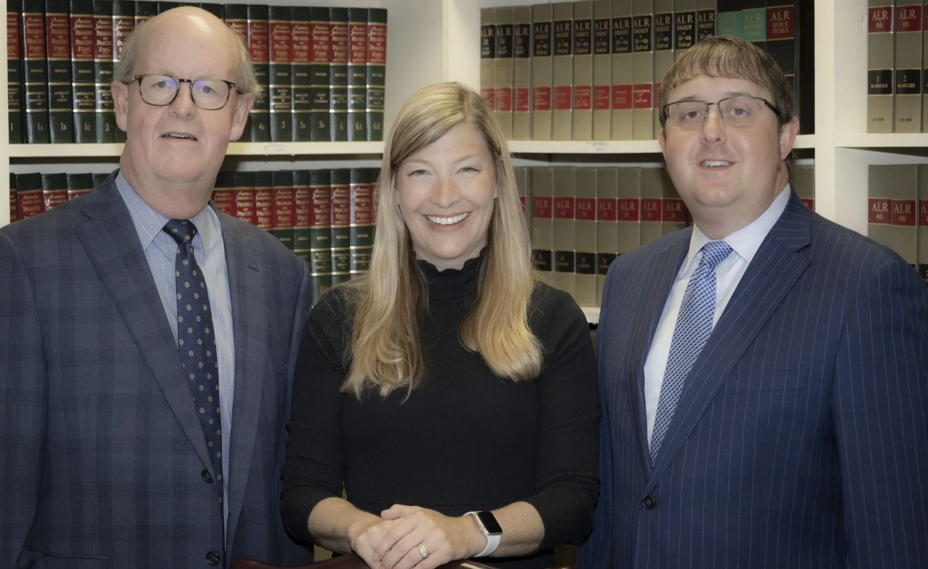 Three people in suits smiling in front of bookshelves, possibly a law office.