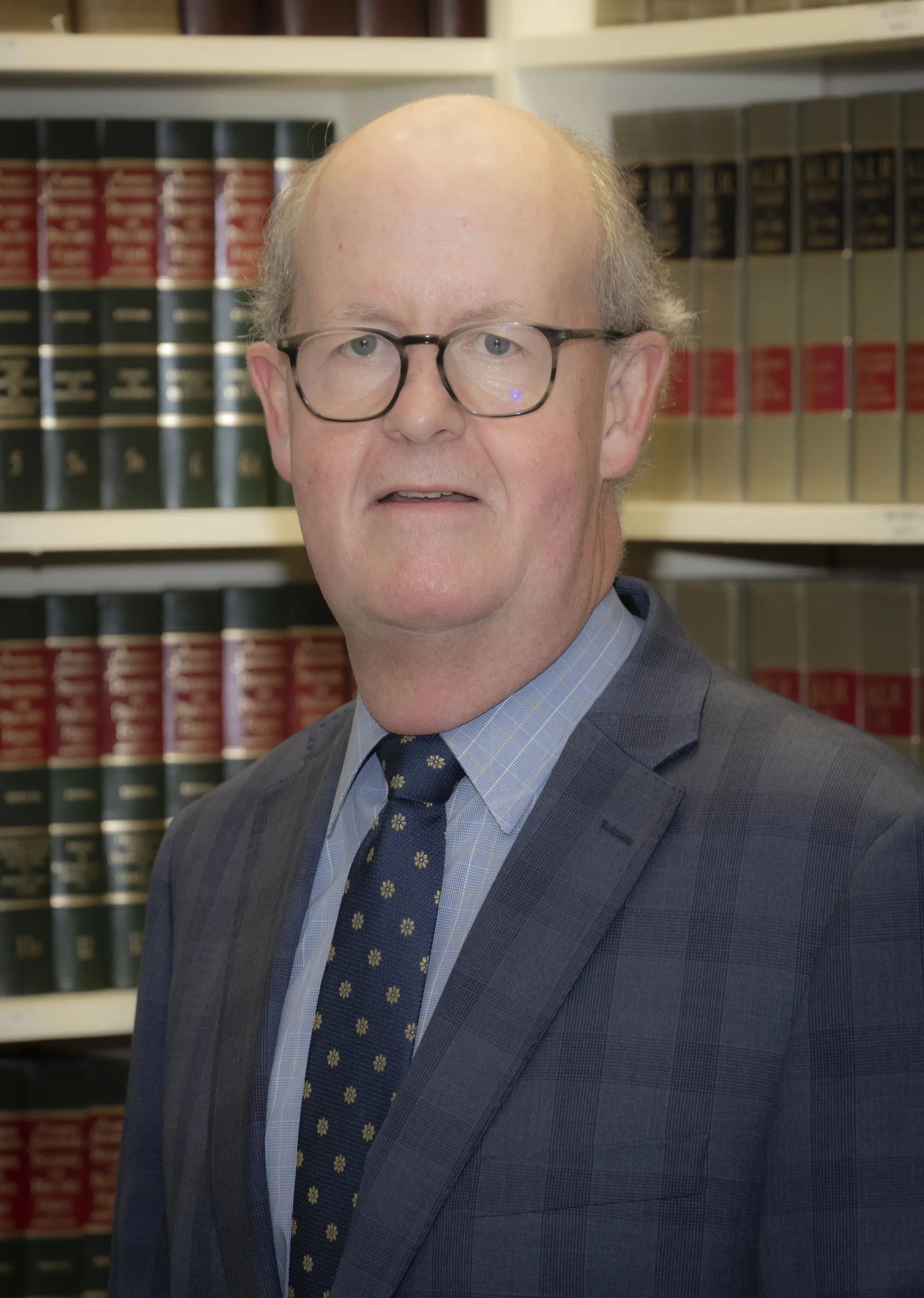 Man wearing glasses, blazer, and tie in front of bookshelves.