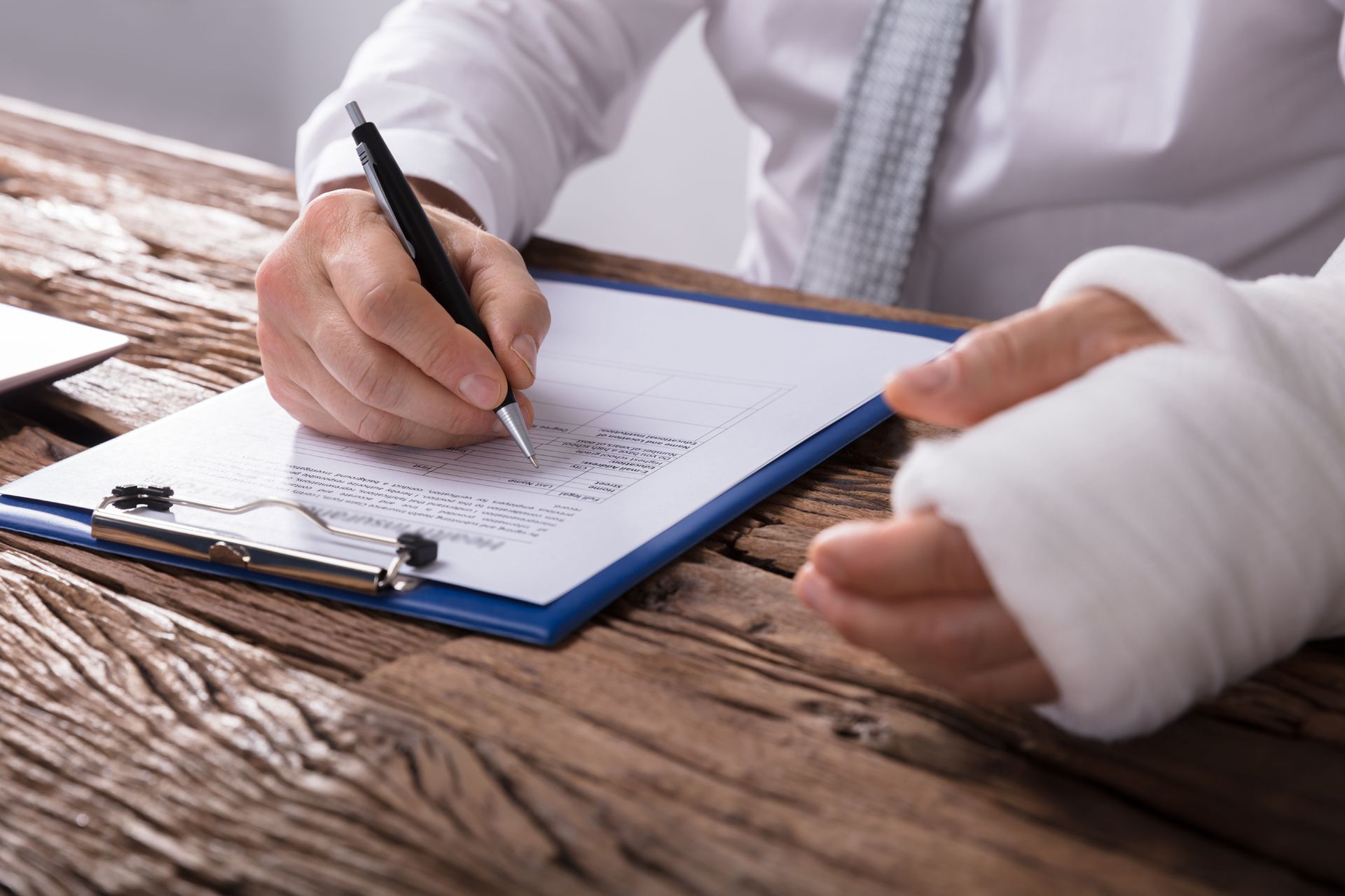 Person with a wrist in a cast writing on a clipboard at a wooden desk.