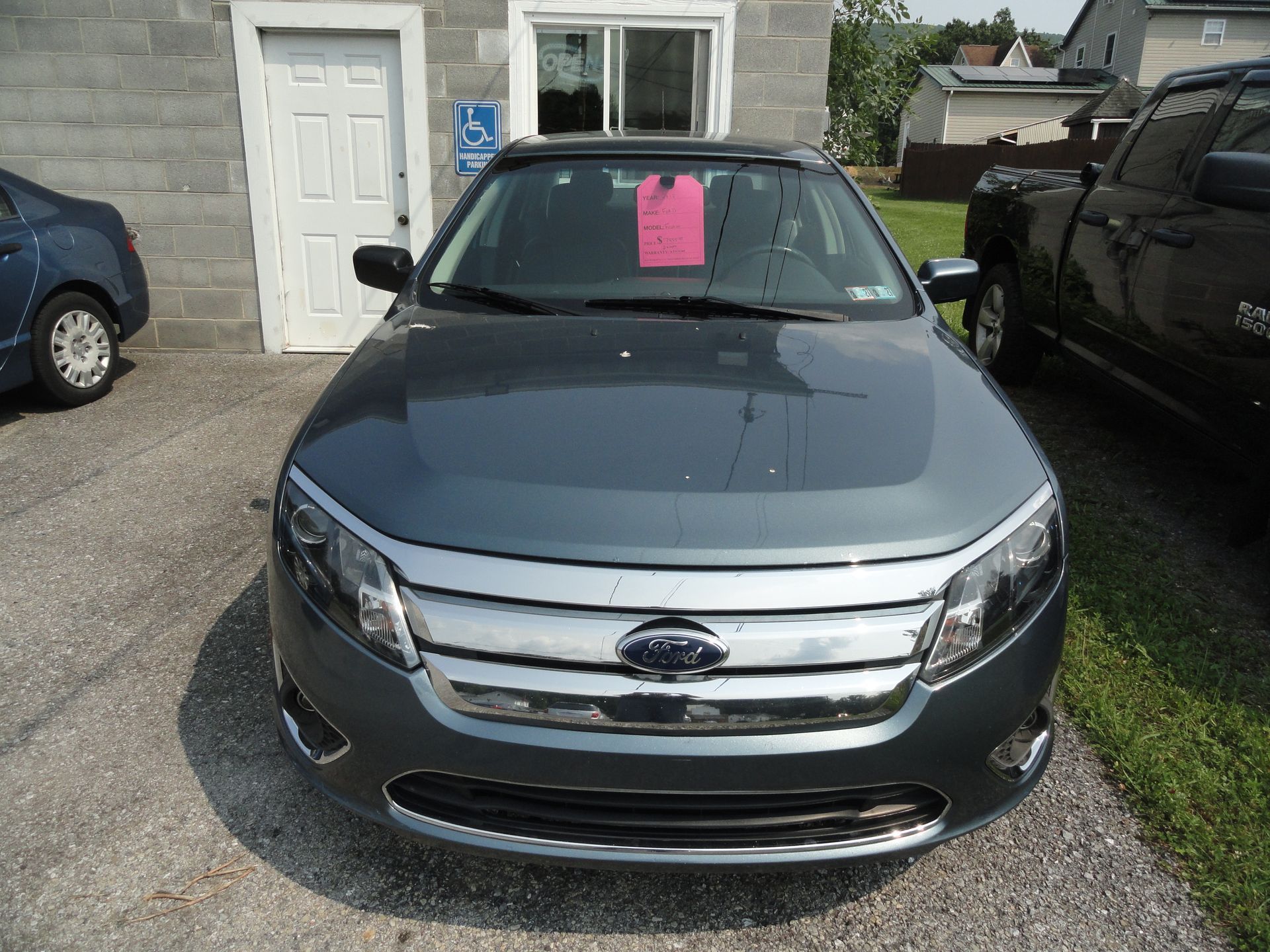 Dark gray Ford sedan parked in front of a house with a pink sale sign in the windshield