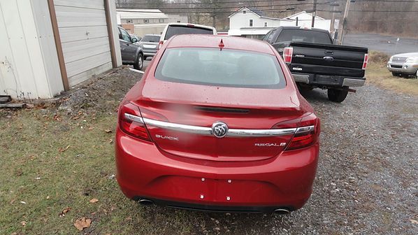 Rear view of a red sedan parked in a gravel lot beside a building, with a black pickup behind it.