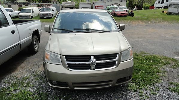Front view of a beige minivan parked on gravel in a lot, with other vehicles in the background