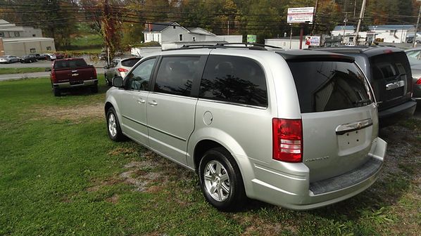 Silver minivan parked on grass near a road with trees and other vehicles in the background
