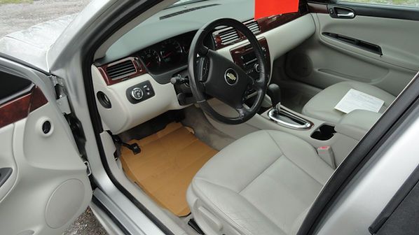 Luxury car interior with white leather seats, black steering wheel, and tan floor mats, viewed from the open door