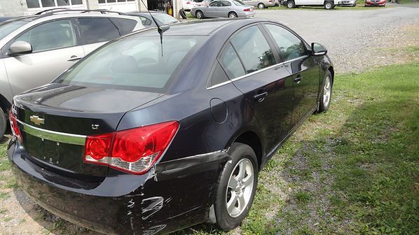 Black sedan parked on grass beside a gravel lot, rear-left view with damaged front fender.